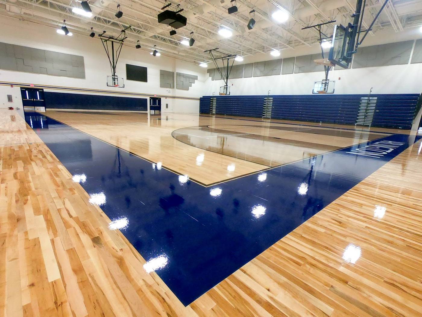 School gymnasium with a glossy maple court, navy accent border, retracted bleachers, and glass backboards