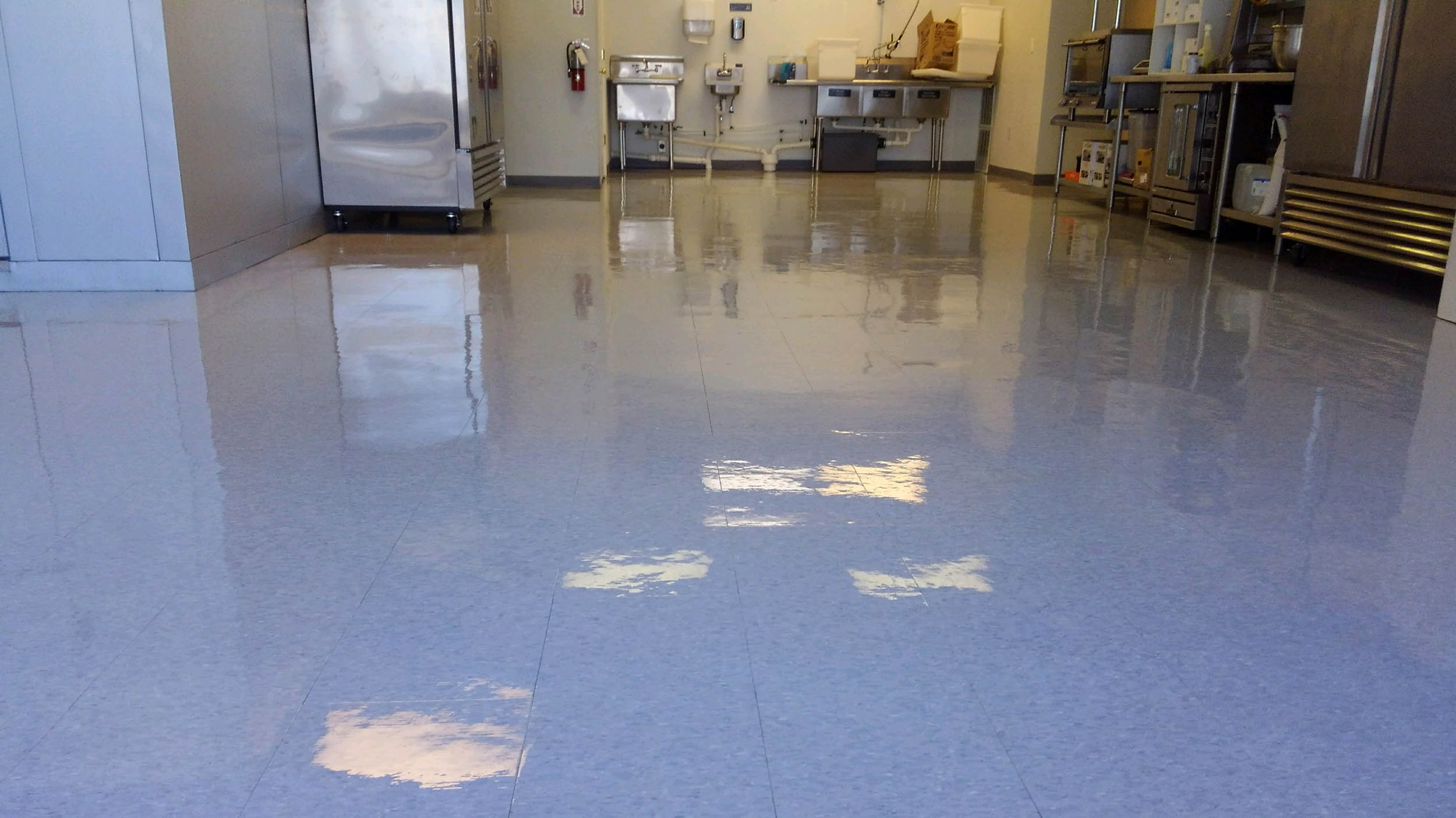 Commercial kitchen with a pale grey speckled VCT floor coated to a high gloss, stainless prep tables and a three-compartment sink along the back wall